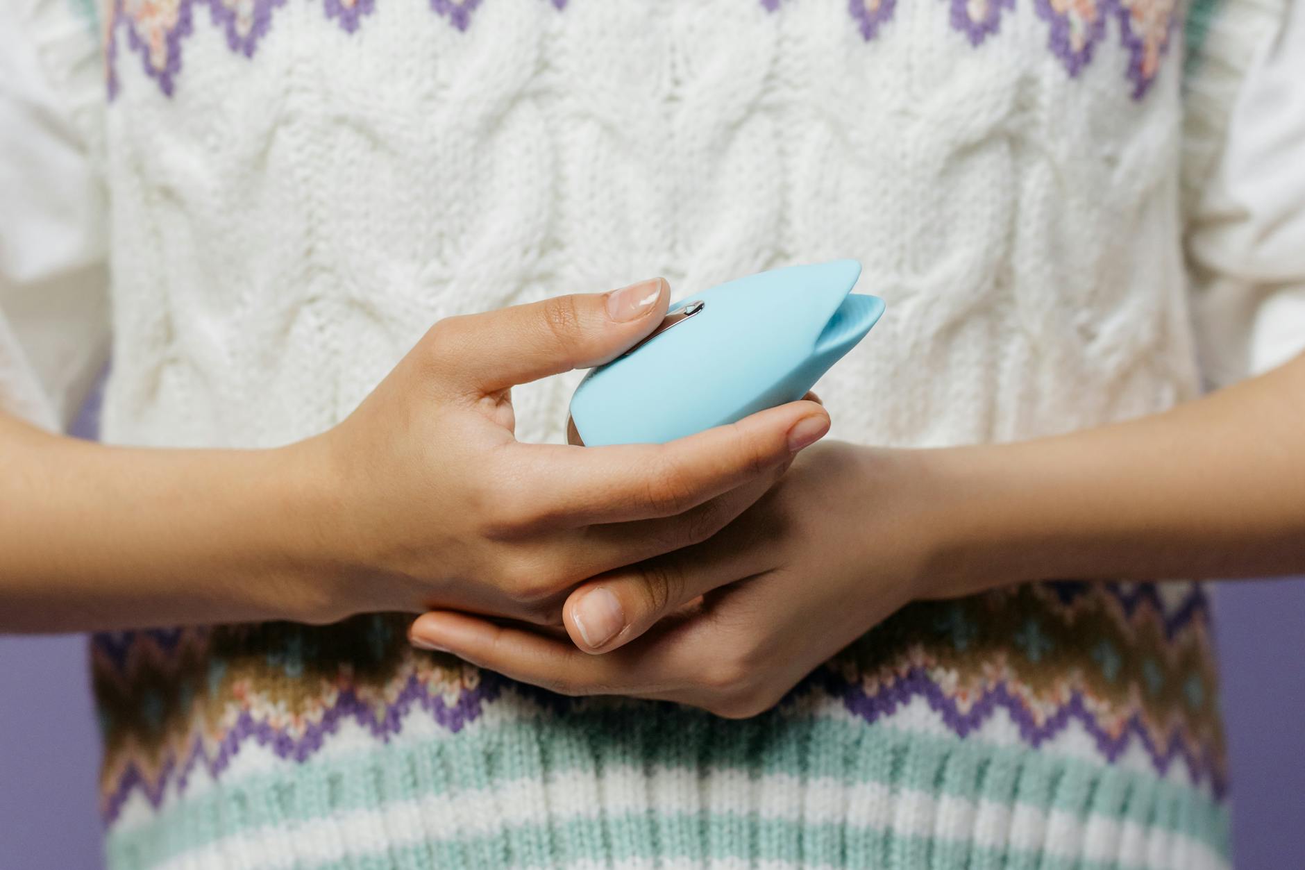 Hands holding a blue personal massager against a knitted sweater, symbolizing intimate self-pleasure.